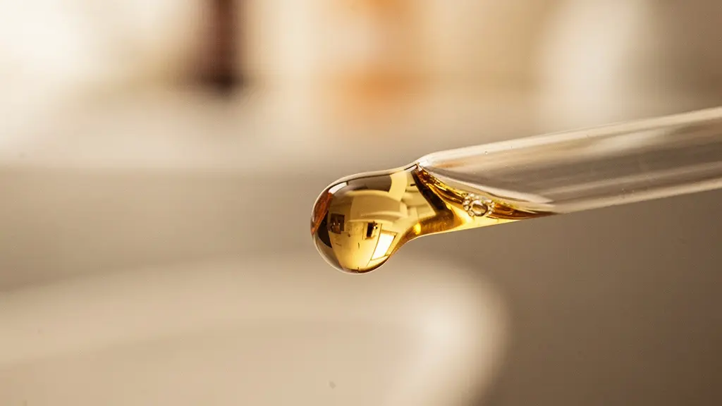 Vue macro d'une goutte d'huile dorée suspendue à l'extrémité d'un flacon compte-gouttes, évoquant le soin auriculaire par approche émolliente