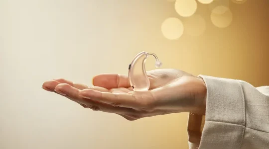 Main ouverte tenant delicatement un appareil auditif miniature translucide dans une lumiere douce et chaleureuse, symbolisant l'accessibilite et la qualite des soins auditifs